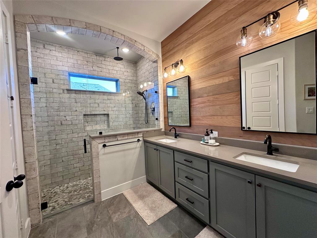 Walk-in shower with brick-tiled walls and arched entry, double vanity with dual undermount sinks, wood-plank wall accent, and matte black hardware