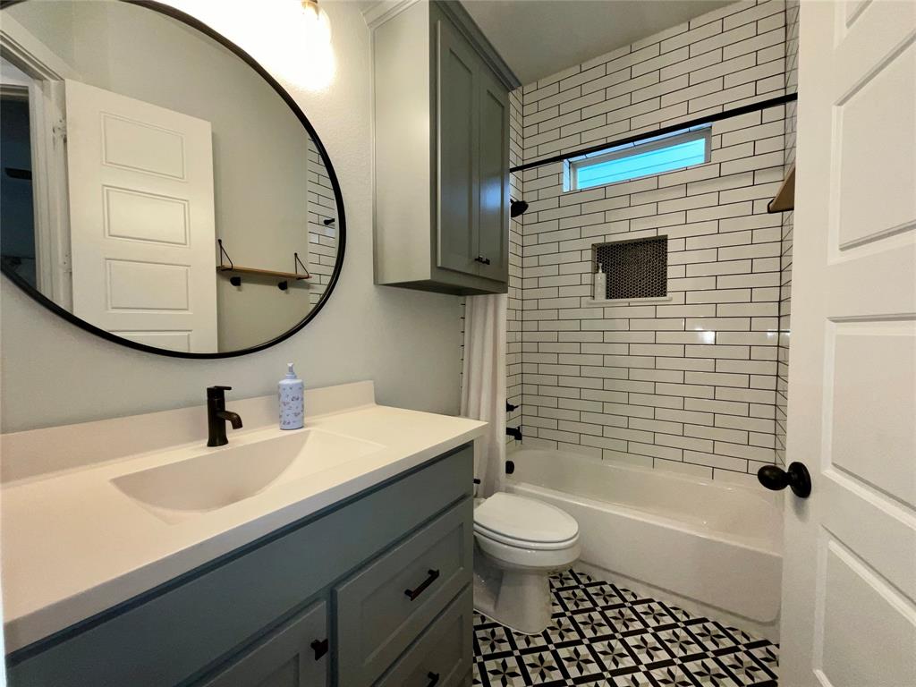 Bathroom featuring a grey vanity with a solid surface countertop, matte black hardware, oval mirror, white subway tile shower surround with a built-in niche, and patterned floor tile