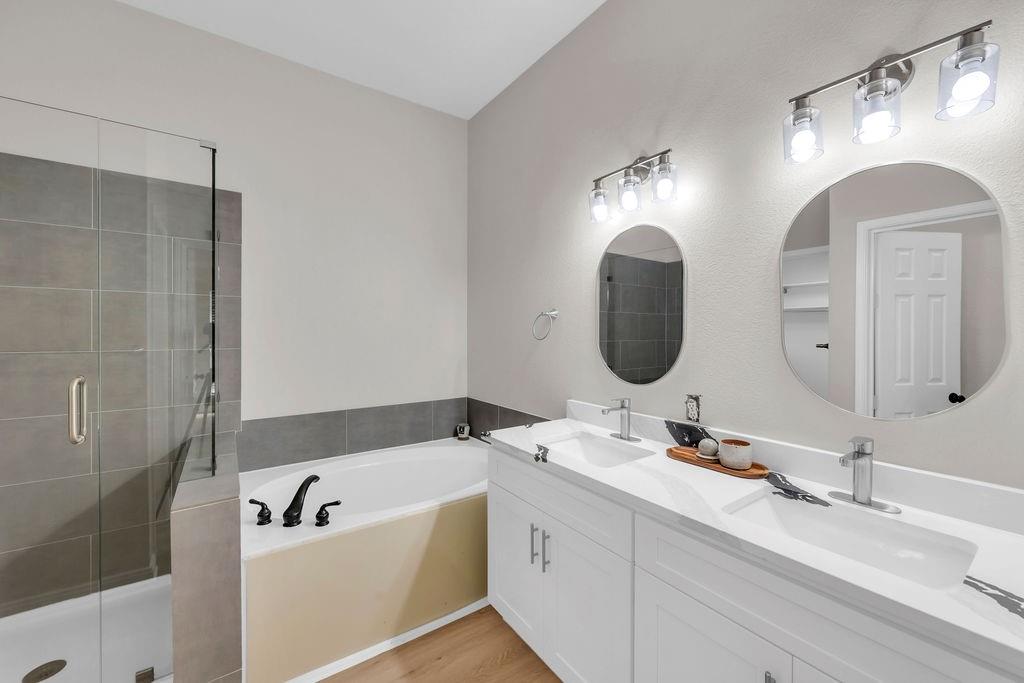 Dual-sink vanity with white cabinetry and solid-surface countertop, walk-in shower with glass enclosure, deep soaking tub, and round mirrors with integrated three-light fixtures