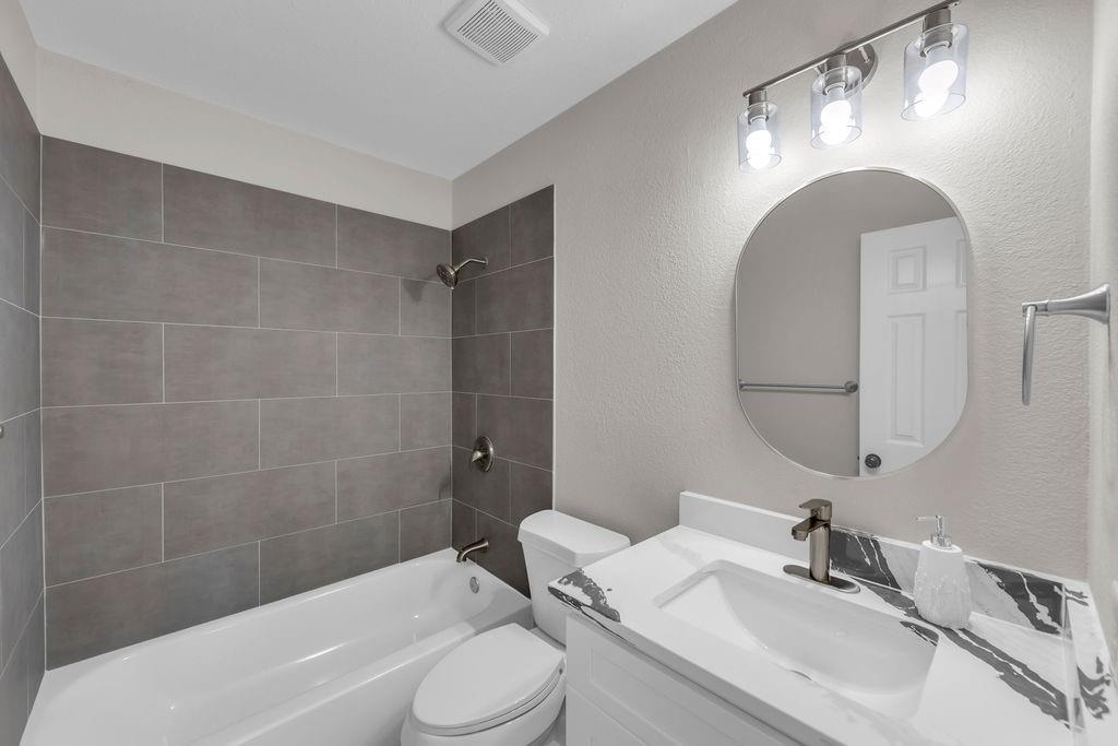 Bathroom featuring a vanity with a patterned countertop, oval wall-mounted mirror, three-light fixture, and a tub-shower combination with floor-to-ceiling gray tile surround