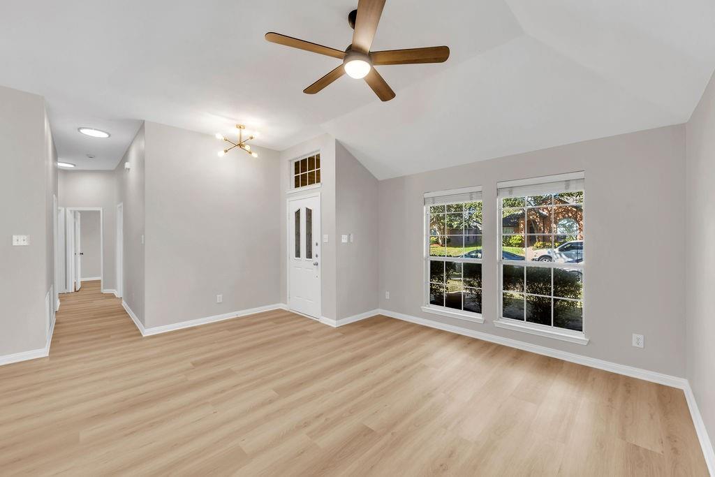Vaulted ceiling featuring a central wood-finish blade ceiling fan, light-toned wood-finish flooring, dual windows with grid inserts, and a white entry door with a glass inlay and transom window