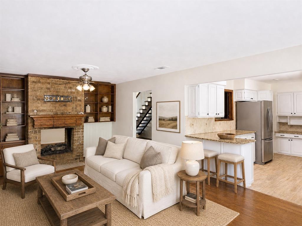 Living area featuring a floor-to-ceiling brick fireplace with wood mantel, built-in wooden shelving, and wood-finish flooring, complemented by an open kitchen with white cabinetry and stone countertops