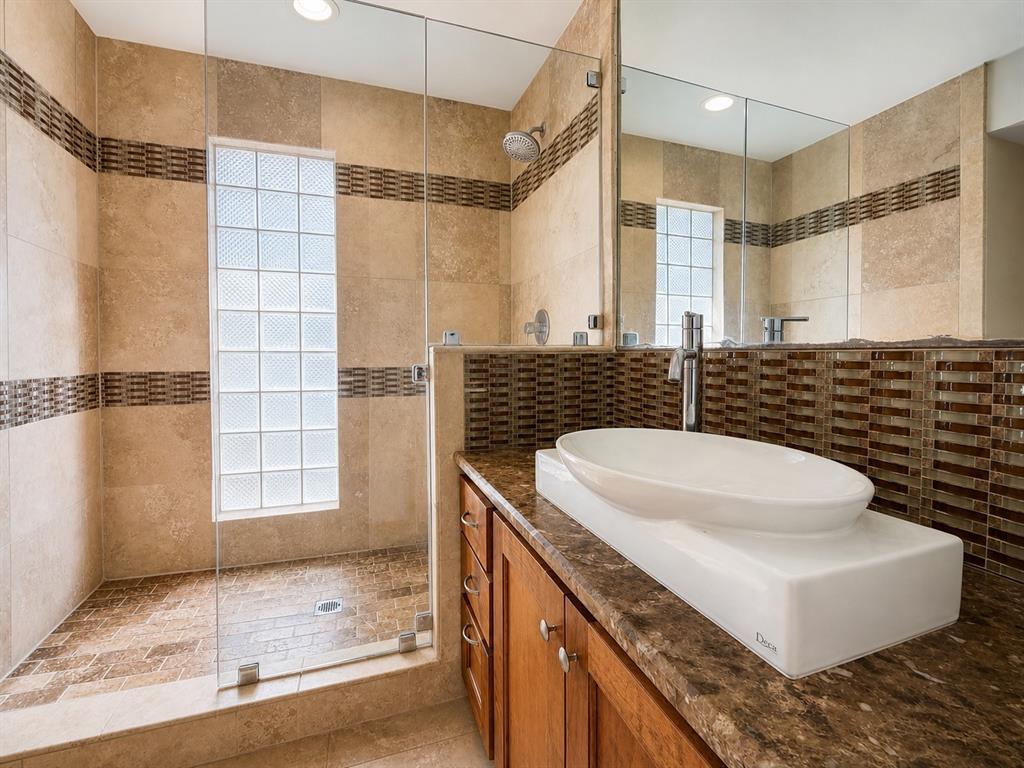Walk-in shower with a glass enclosure, glass block window, stone-finish tile surround, oval vessel sink, and stone-surface vanity countertop