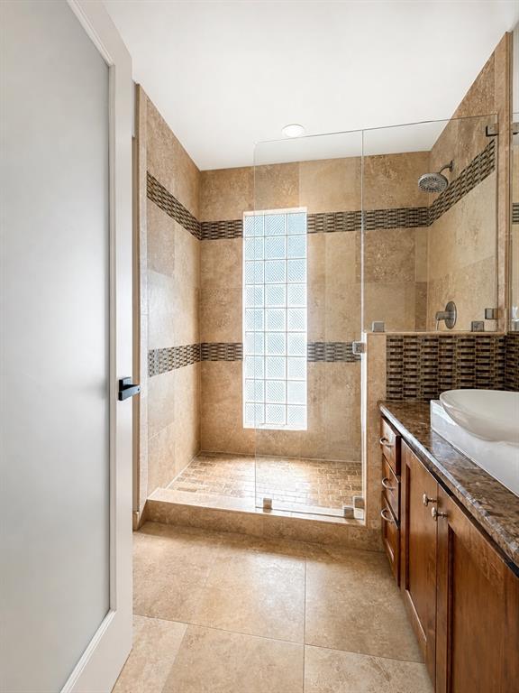 Walk-in shower with glass-block window, stone-tiled walls, glass door enclosure, and wood-finish vanity with a vessel sink and stone countertop