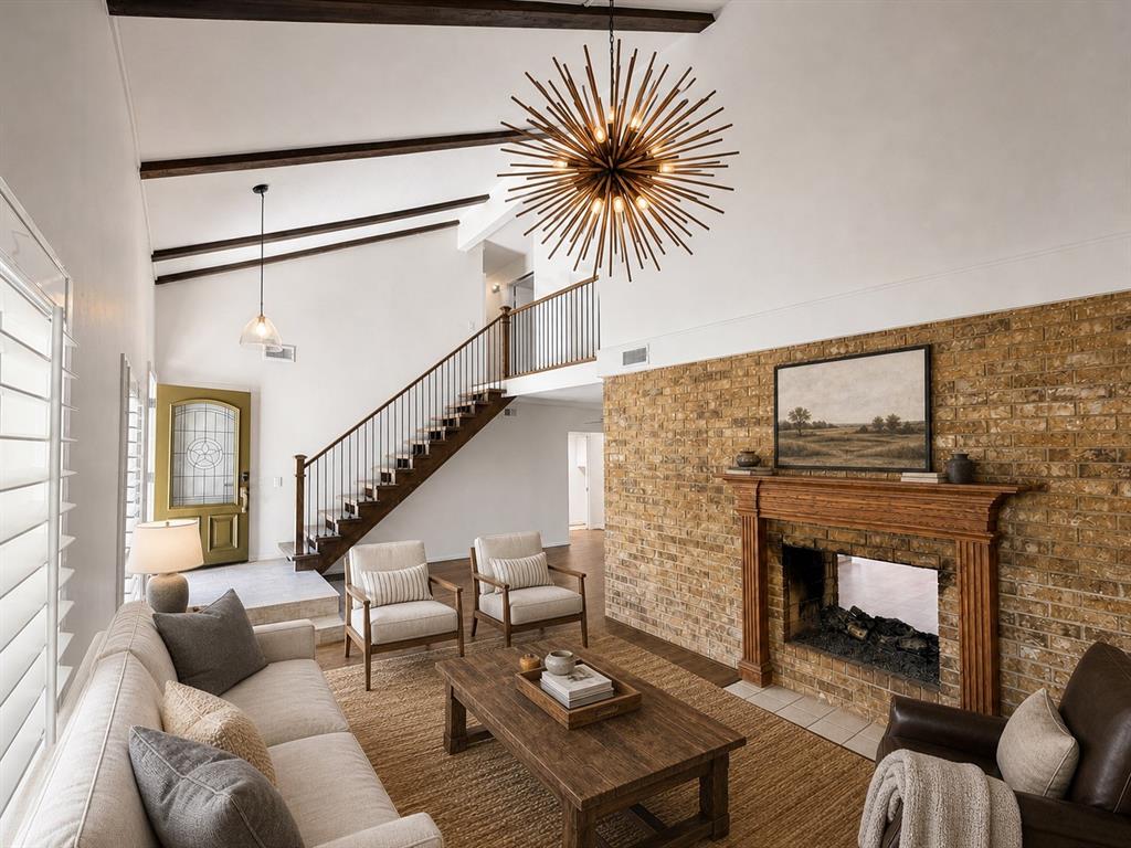 Vaulted ceiling with exposed wood beams, full-height masonry fireplace with carved wood mantel, starburst statement chandelier, and staircase featuring a wood handrail with vertical balusters