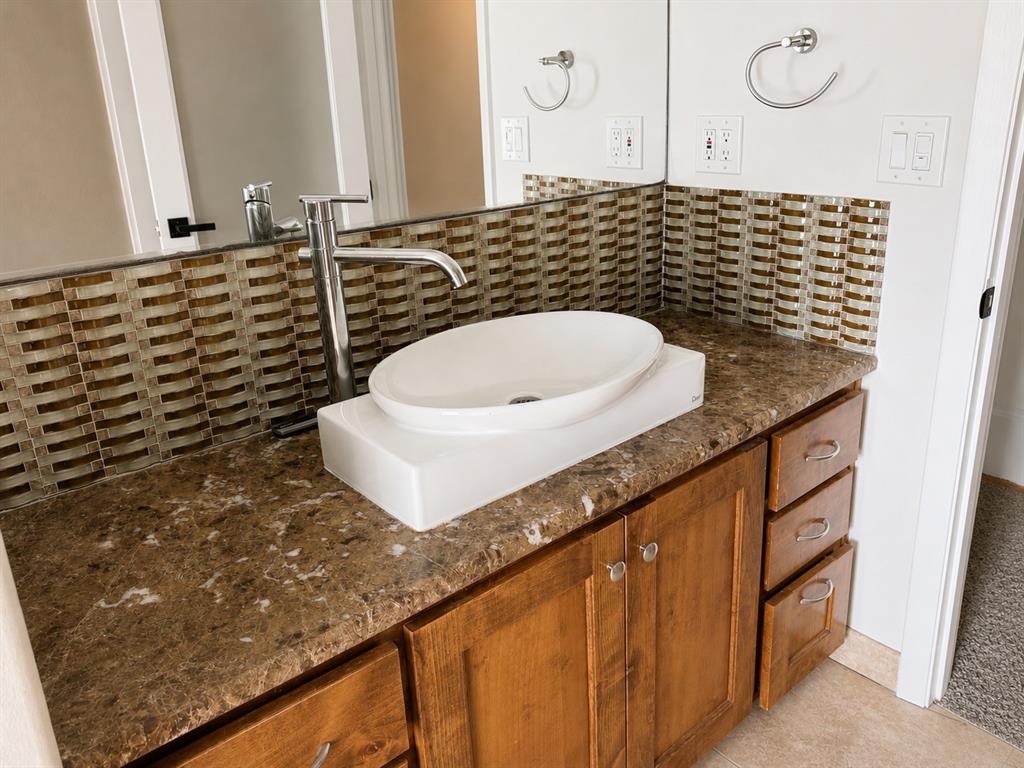 Bathroom vanity featuring a vessel sink, tall metal faucet, stone-finish countertop, wood cabinetry, and a glass tile backsplash