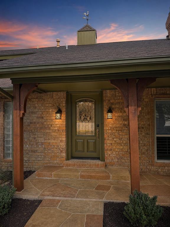 Brick facade entry featuring a flagstone porch, decorative wood support posts, a glass-inset door, and a roof-mounted weathervane Brick facade entry featuring a flagstone porch, decorative wood support posts, a glass-inset door, and a roof-mounted weathervane