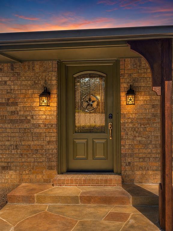 Brick exterior entry featuring a flagstone porch, arched wood-finish front door with decorative glass inlay, and two wall-mounted metal light fixtures