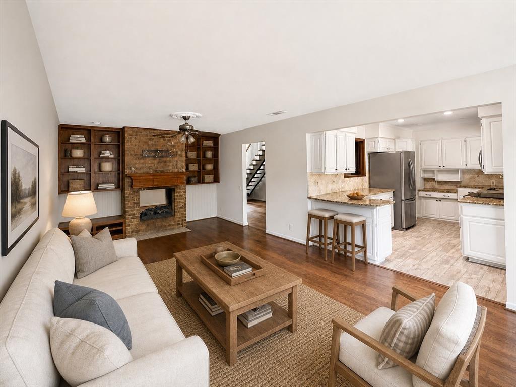 Living area featuring a floor-to-ceiling brick fireplace with wood-finish built-in shelving, wood-finish flooring, and kitchen with white cabinetry and stone countertops