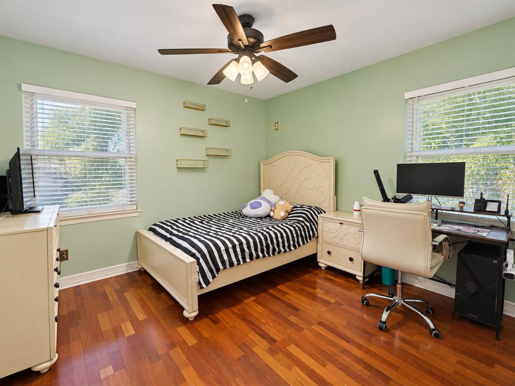 Room featuring wood-finish flooring, two windows with horizontal blinds, built-in wall shelving, and a ceiling fan with integrated lighting