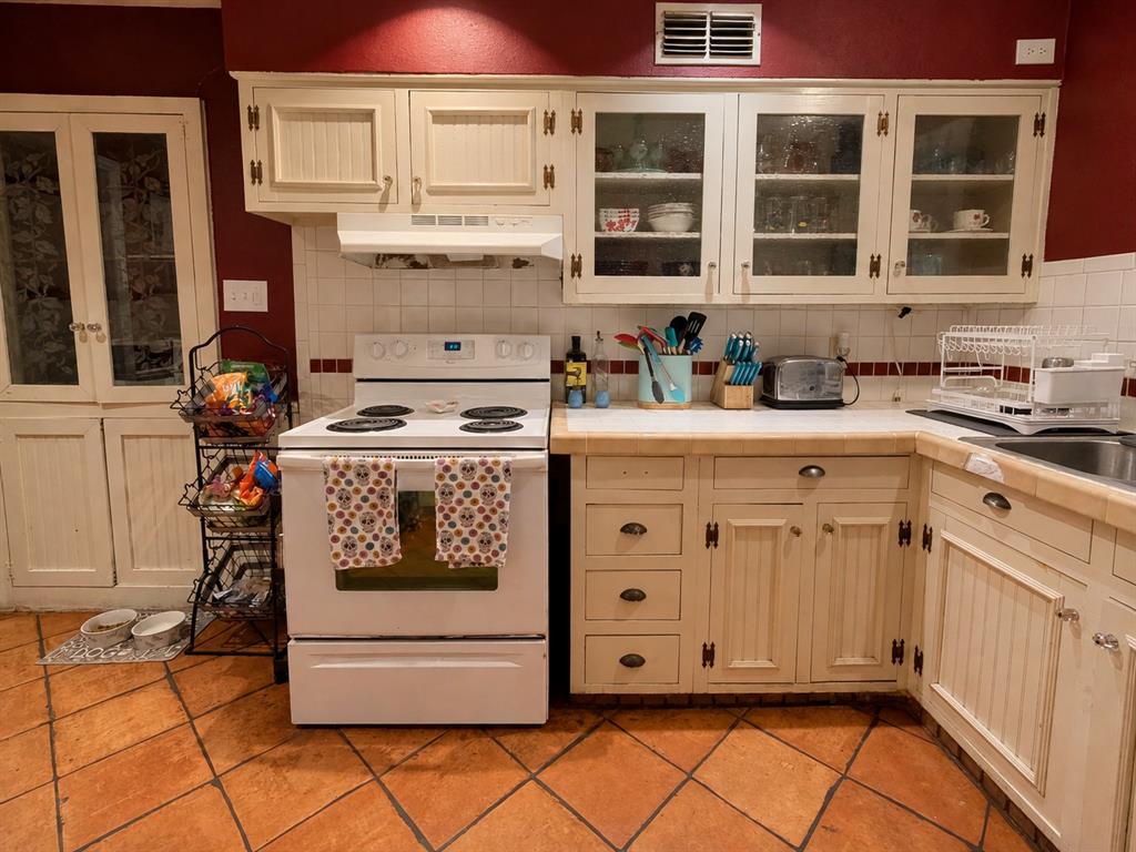 Kitchen featuring off-white cabinetry with glass-front display uppers, tile countertops, terracotta flooring, a built-in pantry, and a white electric range