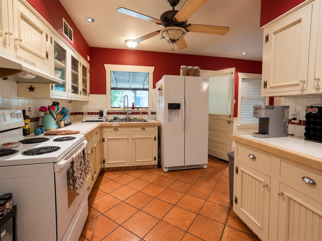 Kitchen featuring cream-colored beadboard cabinetry, terra-cotta tile flooring, tile countertops, glass-front upper cabinet doors, and a ceiling-mounted light fixture with a fan Kitchen featuring cream-colored beadboard cabinetry, terra-cotta tile flooring, tile countertops, glass-front upper cabinet doors, and a ceiling-mounted light fixture with a fan