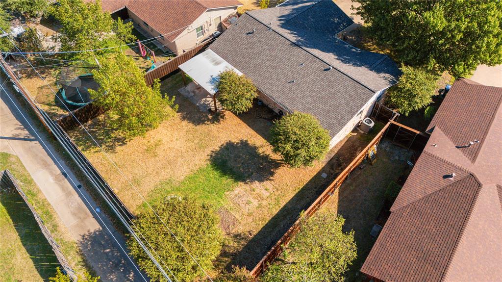 Aerial perspective of a fenced backyard featuring a covered patio, mature trees, and a shingle-roof structure