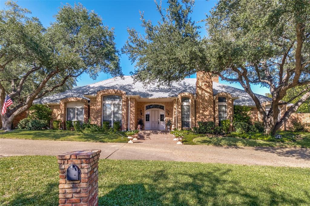 Single-story residence featuring a tan brick exterior, arched window detailing, a chimney, and a concrete driveway bordered by established trees Single-story residence featuring a tan brick exterior, arched window detailing, a chimney, and a concrete driveway bordered by established trees