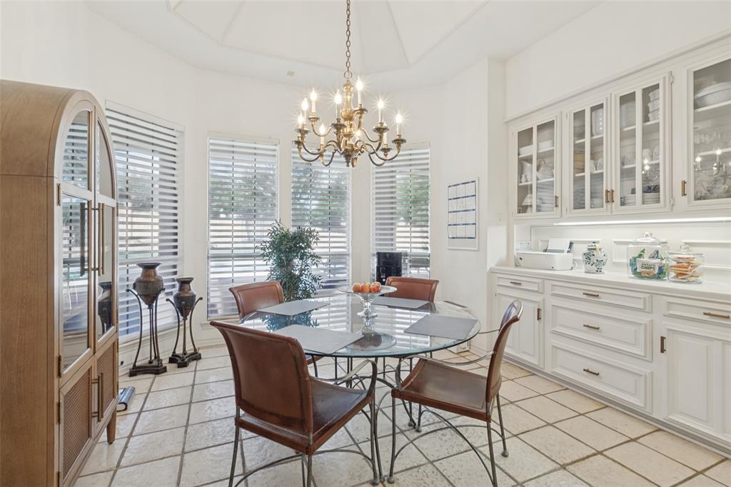 Vaulted ceiling with a multi-arm chandelier, large windows with horizontal blinds, built-in white cabinetry with glass-front display doors, under-cabinet lighting, and light-toned tile flooring