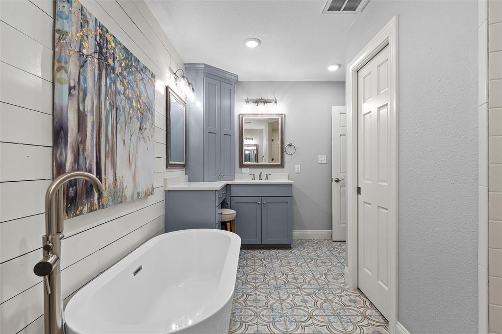 Freestanding soaking tub with a floor-mounted brushed nickel faucet, custom blue cabinetry with integrated tower, patterned tile flooring, and shiplap wall detailing