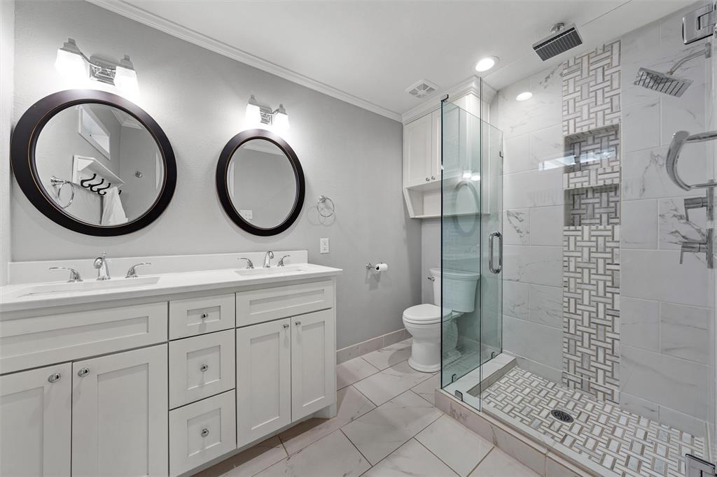 Dual-sink vanity with white cabinetry and countertop, glass-enclosed walk-in shower featuring decorative tile inlay and built-in shelving, and dual circular wall-mounted mirrors