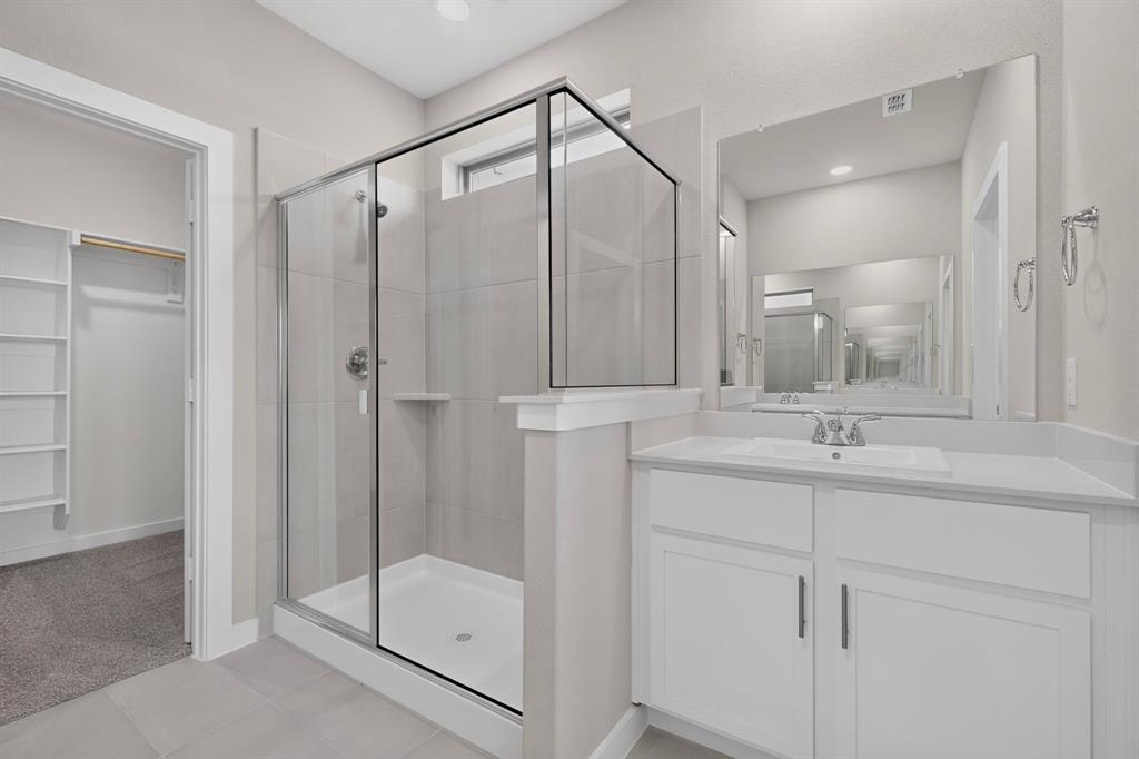 Frameless glass-enclosed shower with tiled surround, white vanity cabinetry with integrated sink, polished hardware, large wall-mounted mirror, and built-in closet shelving