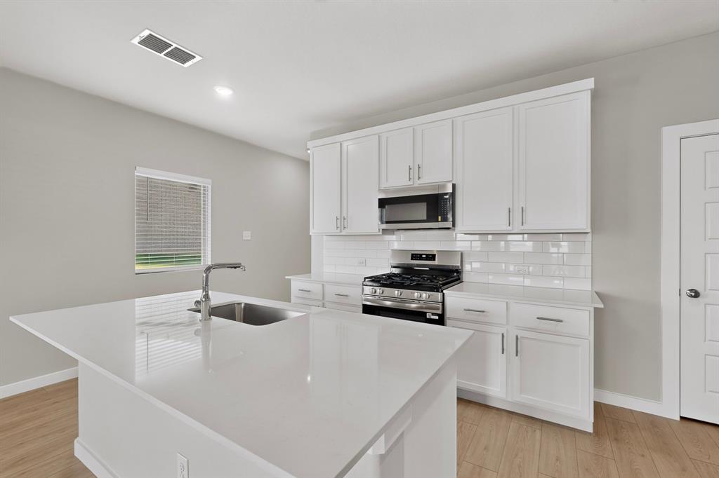 Kitchen featuring white cabinetry, solid-surface countertops, a center island with an integrated stainless steel sink, subway tile backsplash, and wood-finish flooring Kitchen featuring white cabinetry, solid-surface countertops, a center island with an integrated stainless steel sink, subway tile backsplash, and wood-finish flooring