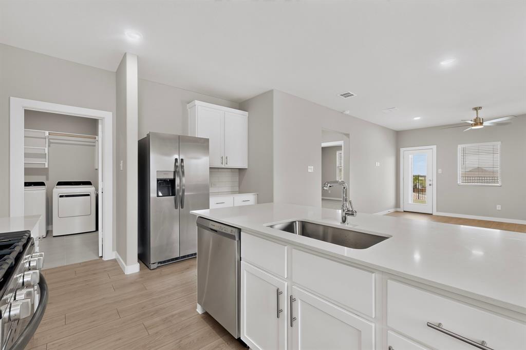 Kitchen featuring an expansive center island with solid-surface countertop, undermount stainless steel sink, white shaker-style cabinetry, and wood-finish flooring Kitchen featuring an expansive center island with solid-surface countertop, undermount stainless steel sink, white shaker-style cabinetry, and wood-finish flooring