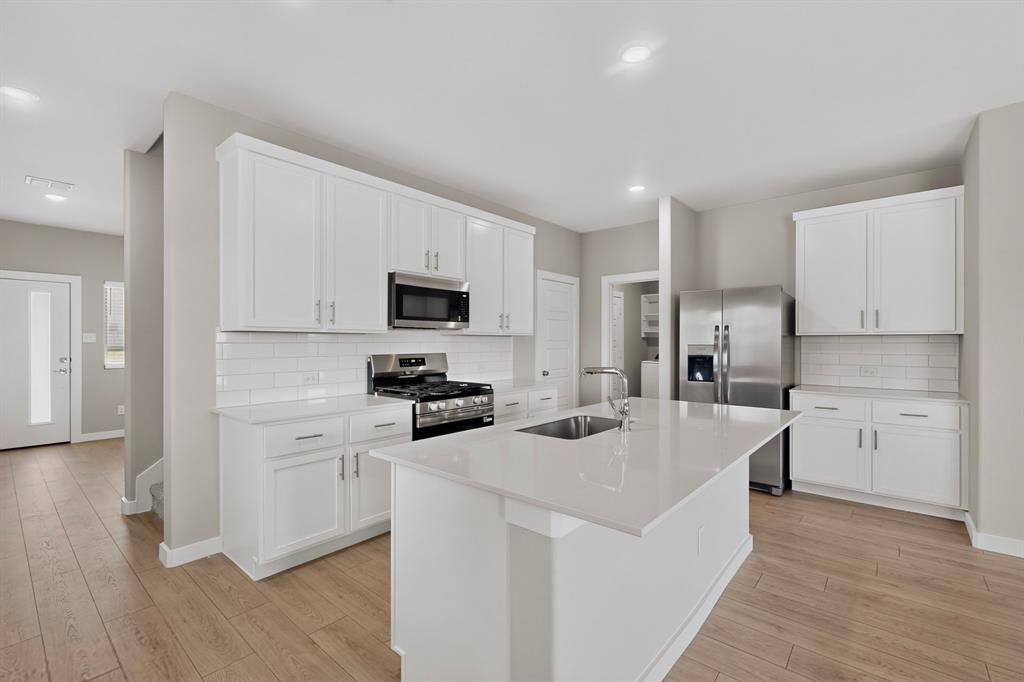 Kitchen featuring white cabinetry, quartz countertops, a central island with a stainless steel undermount sink, subway tile backsplash, and wood-finish flooring Kitchen featuring white cabinetry, quartz countertops, a central island with a stainless steel undermount sink, subway tile backsplash, and wood-finish flooring