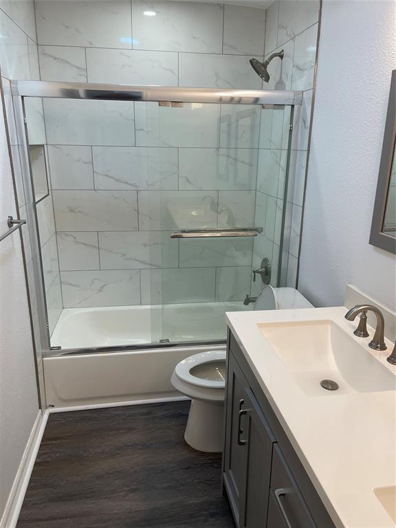 Bathroom featuring wood-finish flooring, a vanity with a solid-surface countertop and undermount sink, shower-tub combination with sliding glass doors, and full-height tiled surround with a built-in recessed niche