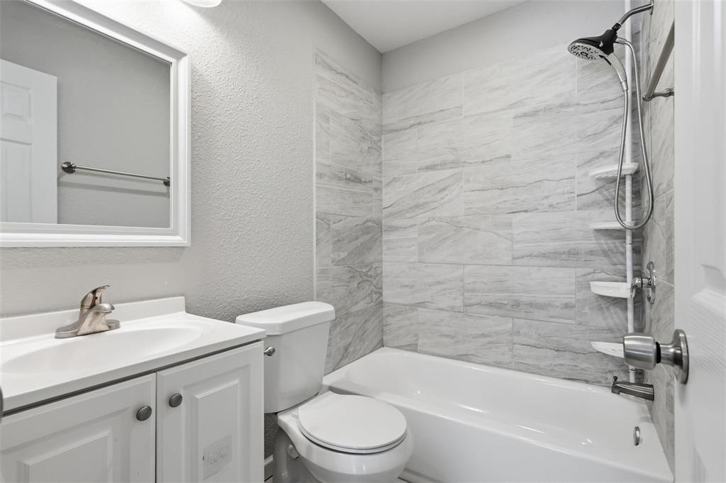 Bathroom featuring a white vanity cabinet, rectangular undermount sink with a polished faucet, wall-mounted mirror, toilet, and a bathtub with stone-patterned tile surround