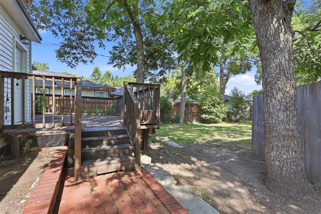Raised wood deck with integrated stairs, red brick pathway, and mature trees providing partial shade within a fenced outdoor area