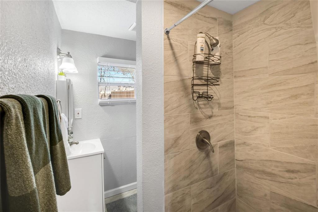 Bathroom featuring a shower stall with neutral-toned, large-format tile surround, single-handle brushed metal faucet, wall-mounted light fixture, and a white cabinet vanity with a built-in sink
