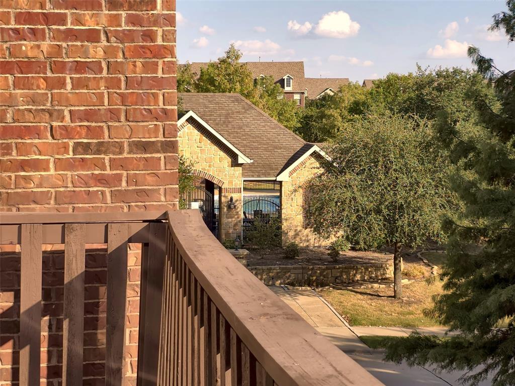 Exterior balcony featuring a wood-style railing and red brick siding, overlooking a neighboring stone-clad building with a shingled roof and mature landscaping