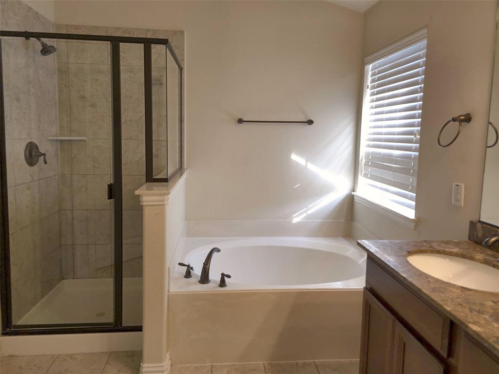 Walk-in shower with stone-finish tile surround and dark metal framing, soaking tub with deck-mounted faucet, wood-finish cabinetry, stone-composite countertop, and under-mount sink