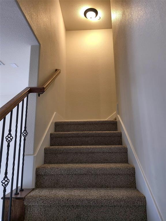 Staircase featuring wall-to-wall carpeting, wood handrail, decorative metal balusters, and a flush-mount ceiling light fixture Staircase featuring wall-to-wall carpeting, wood handrail, decorative metal balusters, and a flush-mount ceiling light fixture