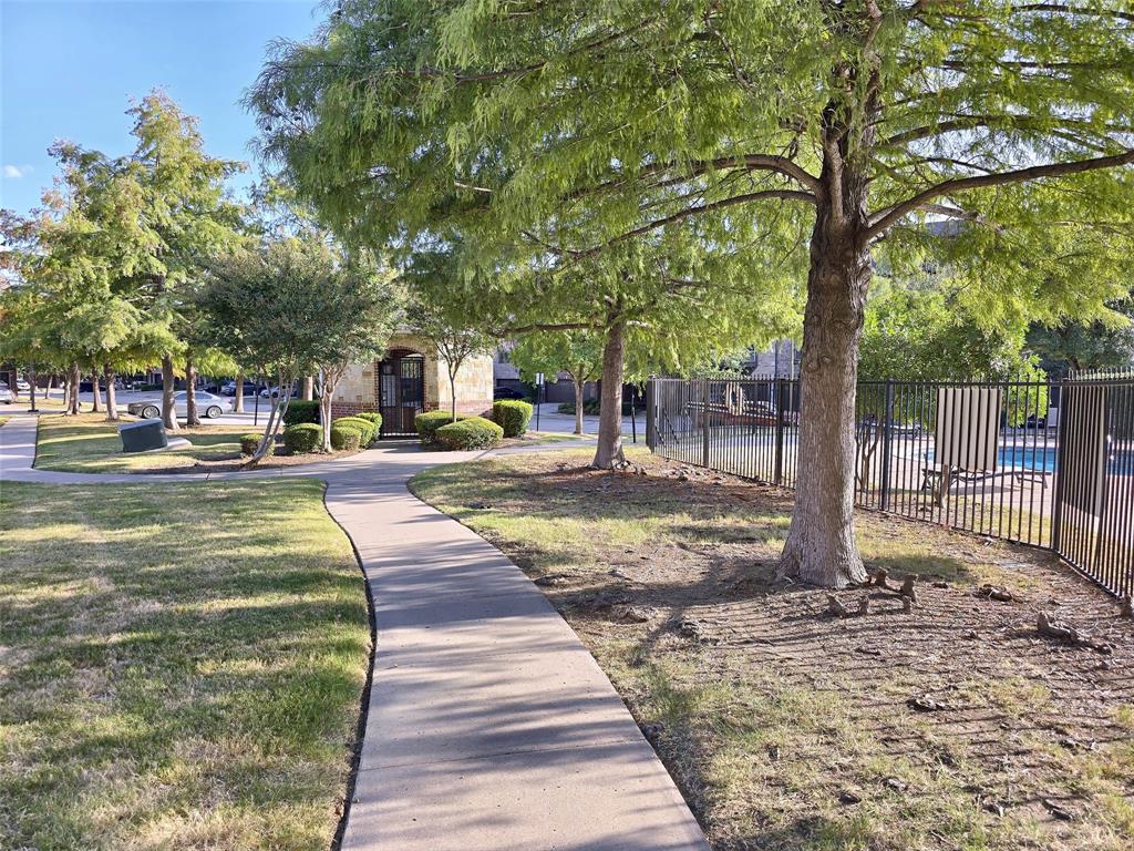 Concrete walking path bordered by mature trees, manicured lawn, established landscaping, and black metal fencing surrounding a pool area