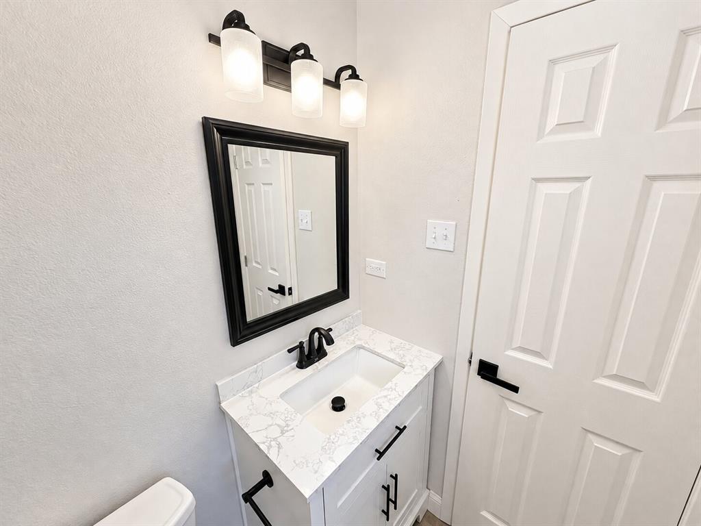 Vanity featuring a white cabinet, stone-finish countertop, and integrated rectangular sink with matte black hardware, accompanied by a framed mirror, three-light fixture with frosted glass shades, and a six-panel door