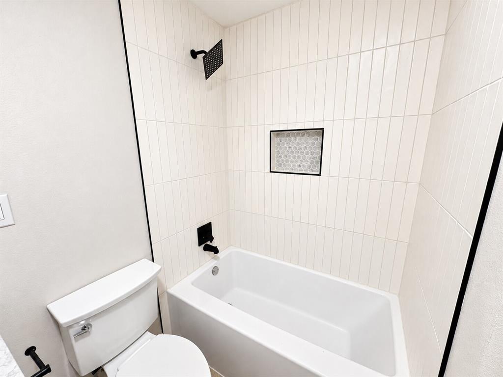 Bathtub alcove featuring vertical white wall tile, matte black shower fixtures, a recessed niche with decorative tile inlay, and a white toilet