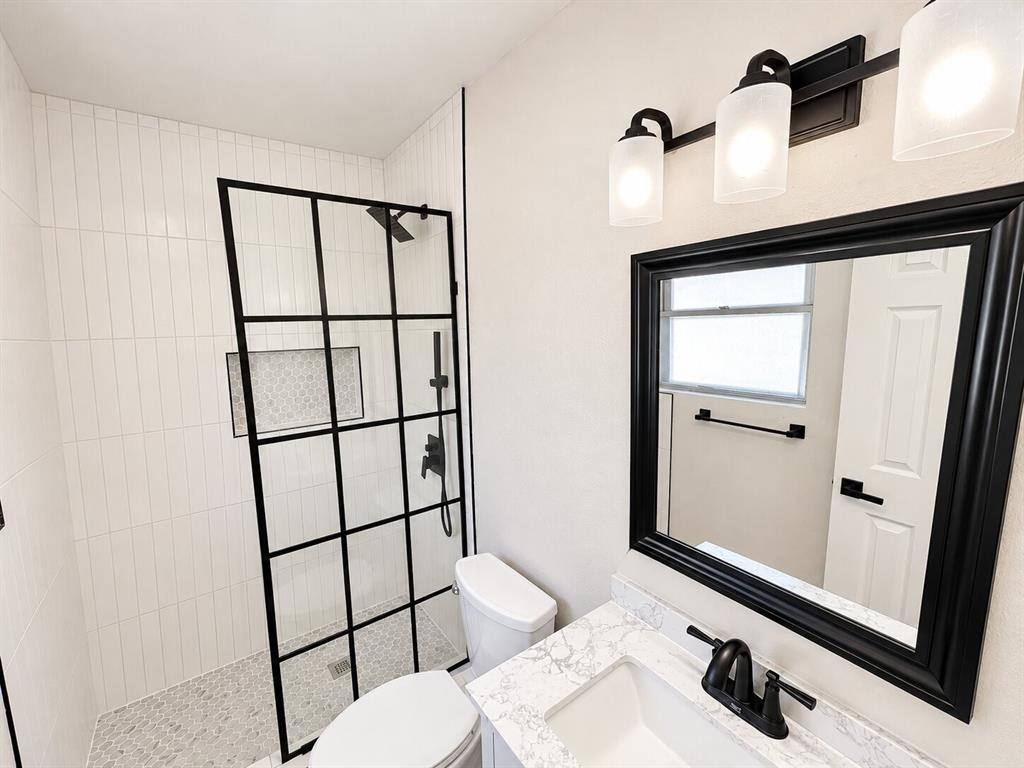 Bathroom featuring a glass shower enclosure with black grid framing, vertical wall tile, stone-patterned countertop, vanity mirror with black frame, and a three-light wall-mounted fixture