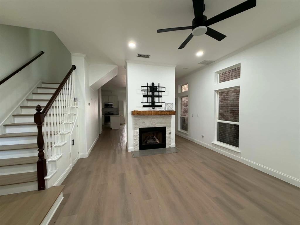 Interior space featuring wood-finish flooring, a central fireplace with stone detailing and a timber mantel, tall windows with brick exterior accents, and a staircase with a dark wood handrail