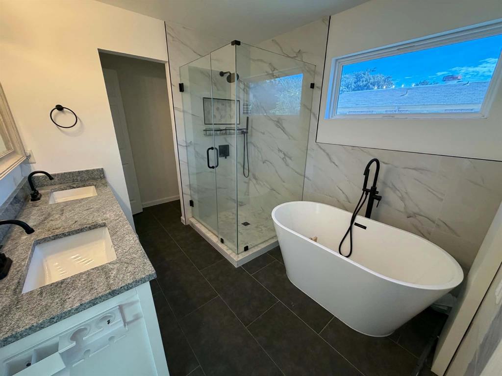 Bathroom featuring a dual-sink vanity with stone countertops, a standalone soaking tub with matte black fixtures, and a glass-enclosed walk-in shower with tile wall surround