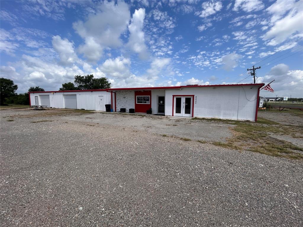 Single-story commercial building featuring white vertical siding, red trim detailing, multiple exterior access points, and a gravel lot Single-story commercial building featuring white vertical siding, red trim detailing, multiple exterior access points, and a gravel lot