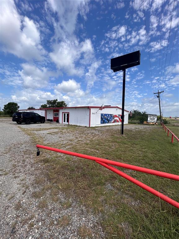 Single-story commercial building featuring white metal siding, a red metal roof, a prominent freestanding electronic signage pylon, and a gated gravel entrance Single-story commercial building featuring white metal siding, a red metal roof, a prominent freestanding electronic signage pylon, and a gated gravel entrance