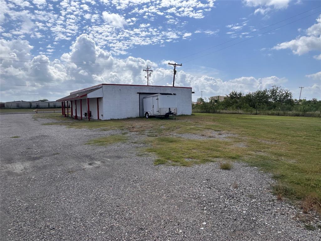 Single-story structure featuring a concrete exterior, a covered side porch with support columns, and an expansive gravel lot Single-story structure featuring a concrete exterior, a covered side porch with support columns, and an expansive gravel lot