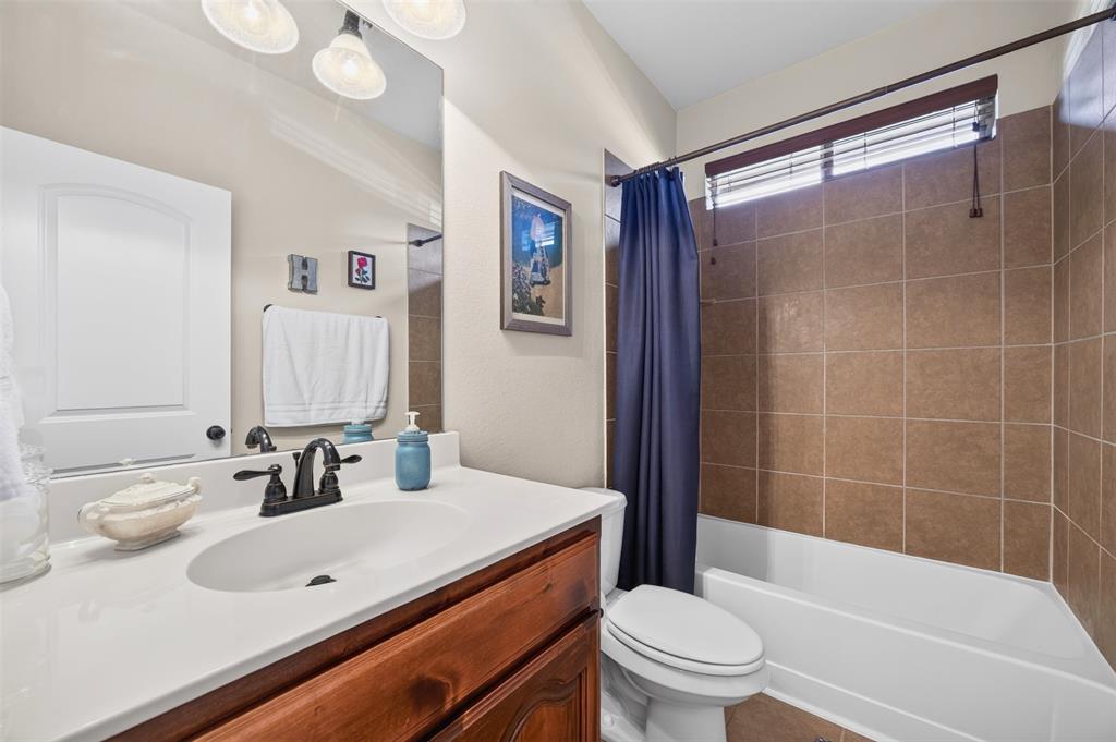Bathroom featuring a wood-finish vanity with a white countertop and oval undermount sink, three-light vanity fixture, and a tub-shower combination enclosed by brown tile surround