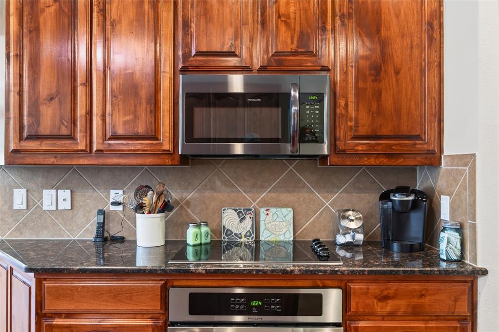 Stained wood cabinetry, stainless steel microwave, dark stone-finish countertop, smooth-top electric cooktop, and diagonal-set tile backsplash