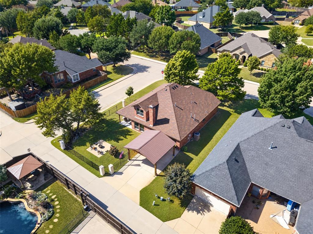 Aerial perspective of a brick residence featuring a metal-roofed carport, a prominent chimney, a fenced perimeter, and established trees on a corner lot