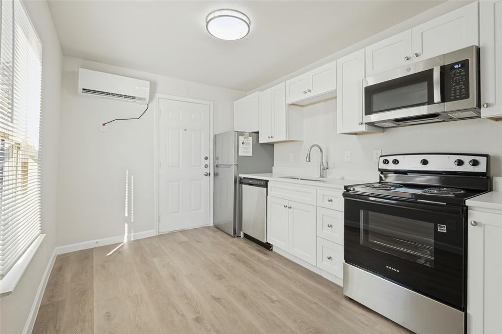 Kitchen featuring white shaker-style cabinetry, wood-finish flooring, stainless steel appliances, and a wall-mounted ductless heating and cooling unit Kitchen featuring white shaker-style cabinetry, wood-finish flooring, stainless steel appliances, and a wall-mounted ductless heating and cooling unit