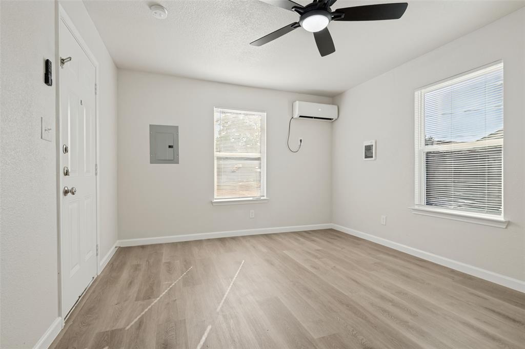 Neutral-toned space featuring wood-finish flooring, a wall-mounted ductless mini-split HVAC unit, two windows with horizontal blinds, and a ceiling fan with integrated lighting Neutral-toned space featuring wood-finish flooring, a wall-mounted ductless mini-split HVAC unit, two windows with horizontal blinds, and a ceiling fan with integrated lighting