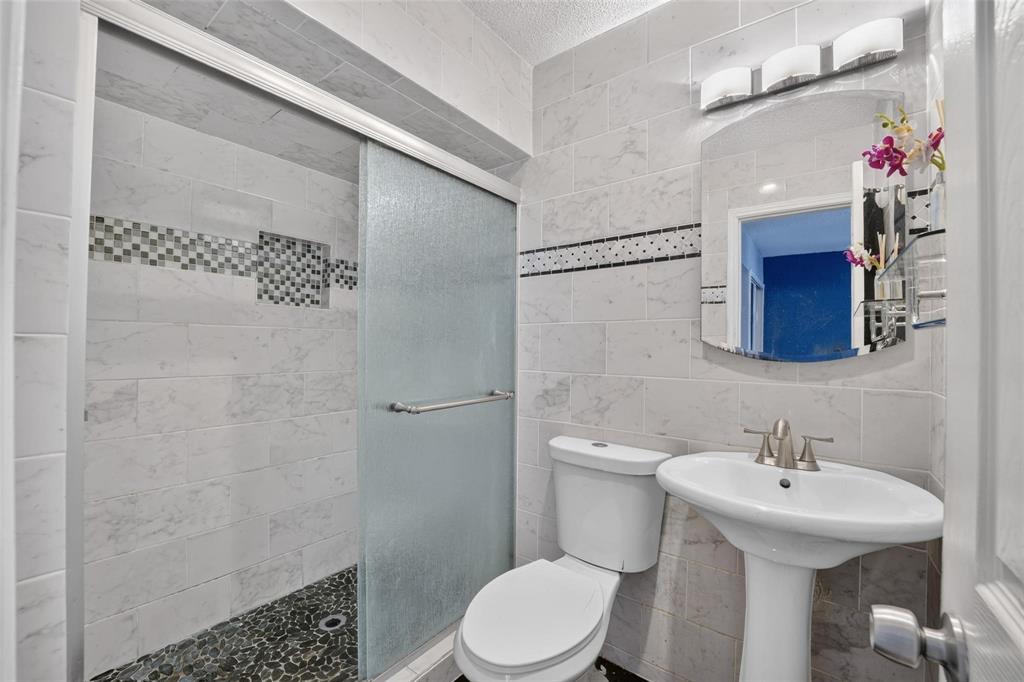 Full bathroom featuring a shower with a sliding glass door, stone-finish tile wall surround, pebble-style flooring, a pedestal sink, and a three-bulb wall-mounted light fixture