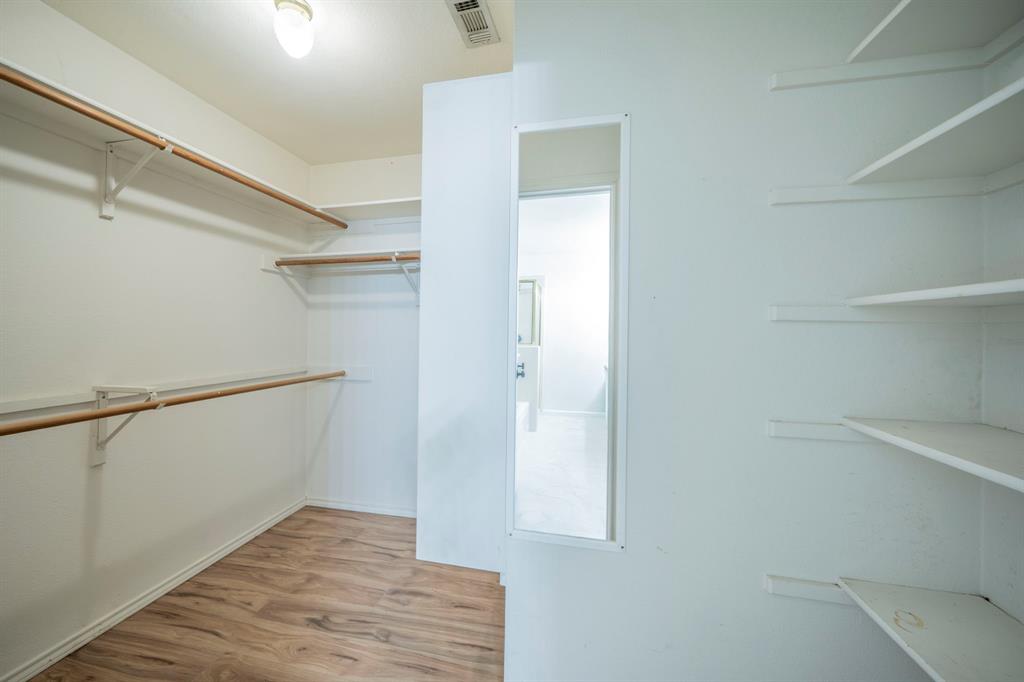 Walk-in storage space featuring wood-finish flooring, white built-in shelving, hanging rods, and a full-length wall-mounted mirror