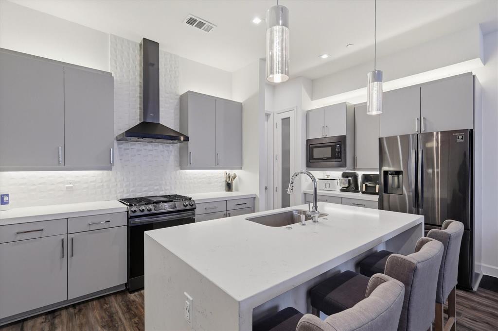 Modern kitchen featuring grey flat-panel cabinetry, quartz countertops, a central island with an integrated sink, textured tile backsplash, and stainless steel appliances