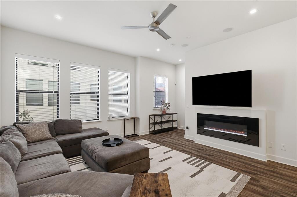 Living area featuring wood-finish flooring, a wall-mounted electric fireplace, recessed lighting, a modern ceiling fan, and large windows with horizontal blinds